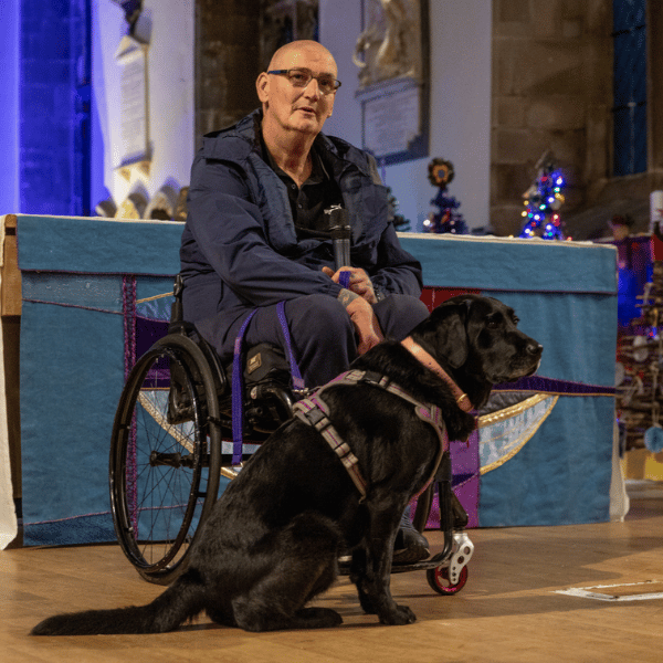 Man in a wheelchair with his assistance dog. Man is holding a microphone.
