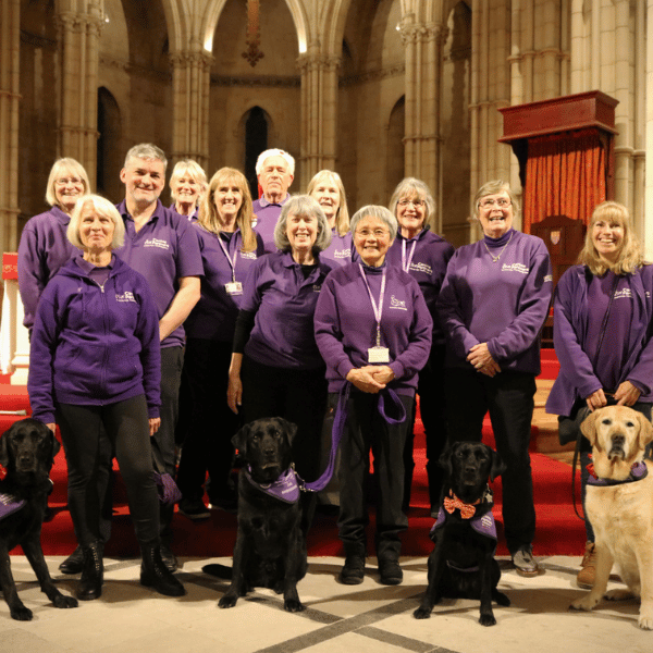A group of people smile at the camera wearing their canine partners jumpers, with a few ambassador dogs sat in front