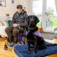 Photo of man in wheelchair in his front room with his Canine partner in a jacket on her bed