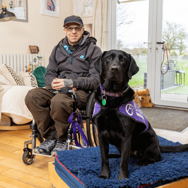 Photo of man sat in wheelchair with black Labrador sat on its bed