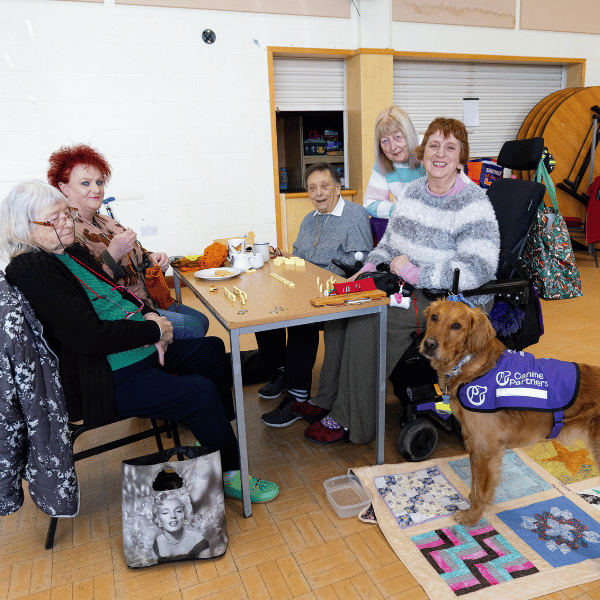 Photo of group of people playing board games with lady in wheelchair and her assistance dog by her side in her purple jacket
