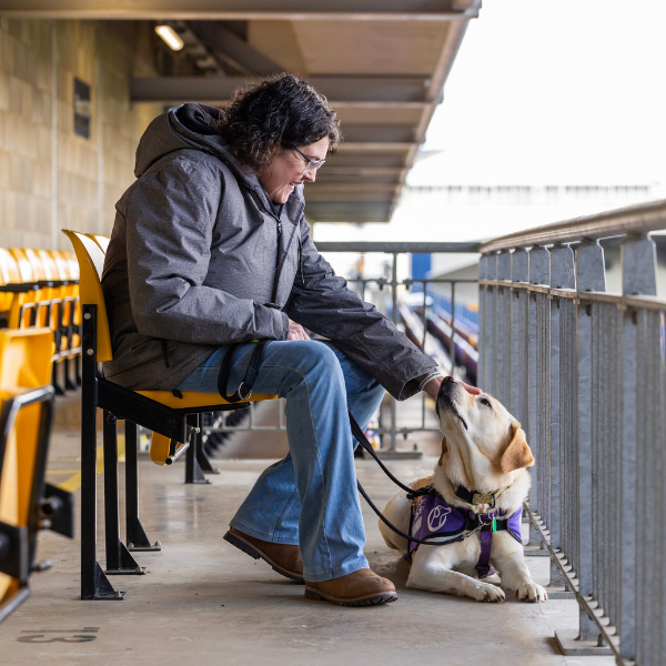 Photo of lady sat on a football stadium seat with her yellow Canine partner in front of her on the floor in a jacket