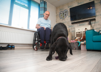 Photo of man in living room sat in his wheelchair with black dog picking up a phone